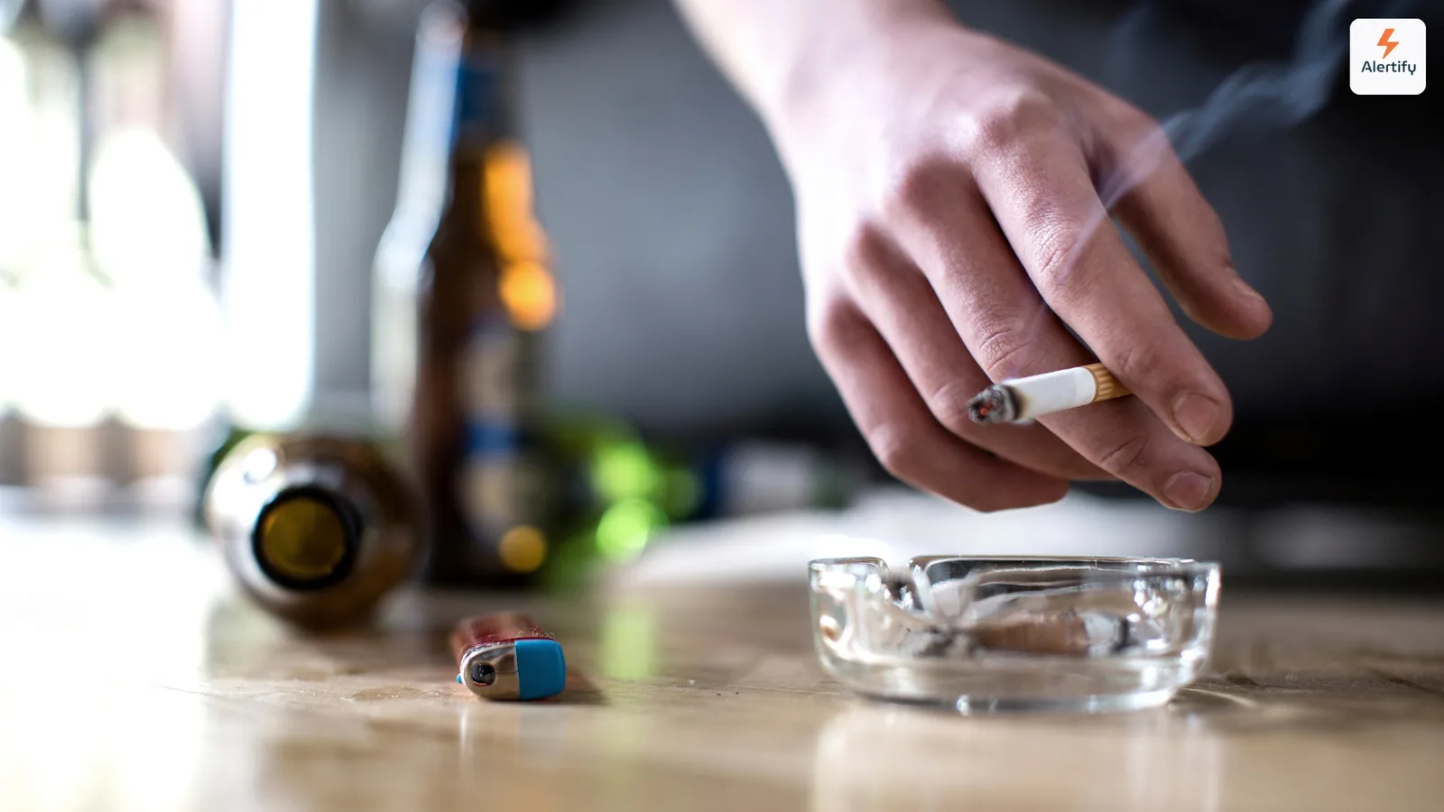 Voyageur tenant une cigarette allumée au-dessus d'un cendrier en verre, à côté de bouteilles vides sur une table
