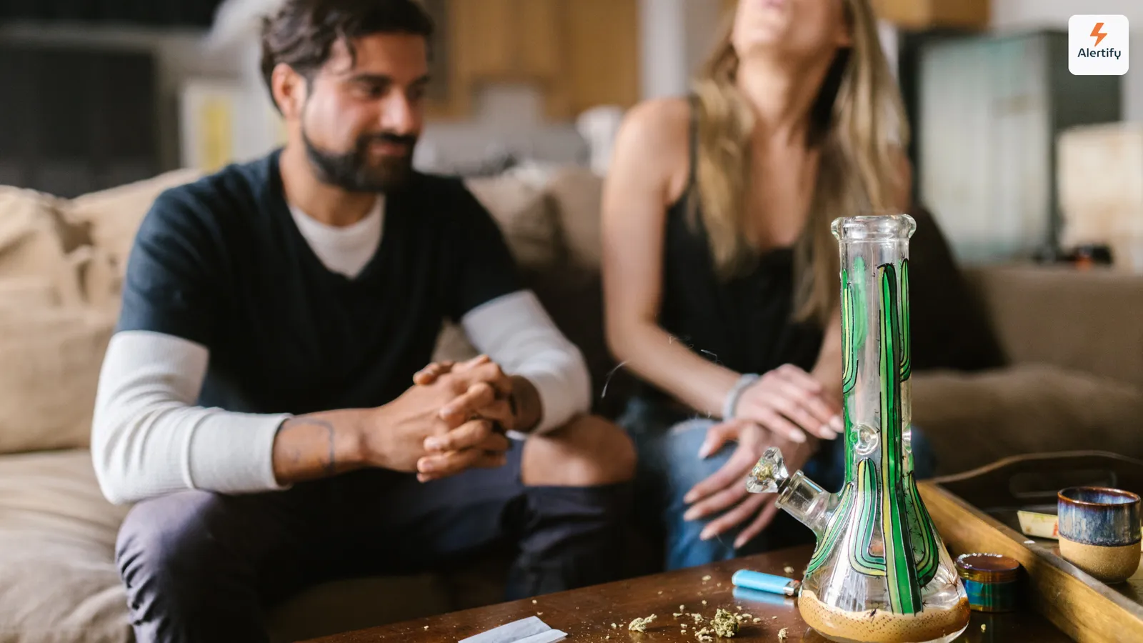 Deux voyageurs assis sur un canapé dans le salon d'une location, avec un bang en verre, un briquet et un grinder sur la table basse devant eux.