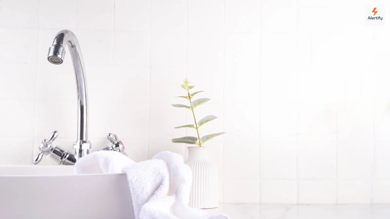 Une salle de bain lumineuse aux murs carrelés blancs, avec un lavabo, une serviette pliée et une petite plante en pot — le type de pièce humide où la moisissure s'installe en premier.